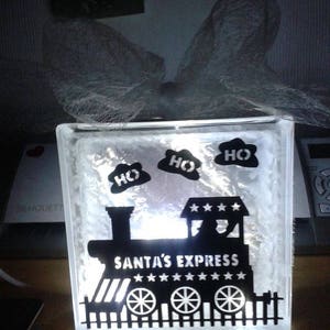 May include: A white glass block with a black silhouette of a train with the words "Santa's Express" and three "HO HO HO"s above the train. The block is lit from inside, making the silhouette glow.