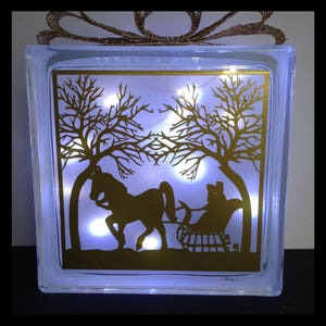 May include: A glass block featuring a gold silhouette of a horse-drawn sleigh and trees. The block is lit from behind, producing a warm glow.