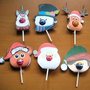 Könnte beinhalten: Sechs Weihnachts-Lollihalter mit verschiedenen Figuren: ein Einhorn, ein Schneemann, ein Rentier, ein Bär, der Weihnachtsmann und ein Elf. Jeder Halter hat ein Loch für einen Lolli und ein festliches Design.