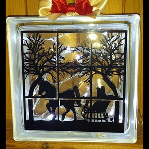 May include: A glass block with a black silhouette of a horse-drawn sleigh with trees in the background. The block is illuminated with white lights.