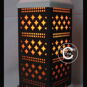 Puede incluir: Una linterna de papel blanca con un diseño geométrico negro y luz naranja que brilla a través. La linterna tiene una base cuadrada y una parte superior puntiaguda. El diseño presenta un patrón repetitivo de diamantes y puntos.