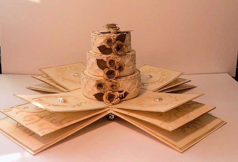 Op de afbeelding: Een drielaagse bruidstaart gemaakt van papier met een kantontwerp en bruine papieren rozen. De taart staat op een witte, gevouwen kaart met een gouden rand.