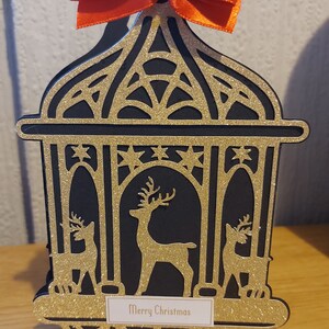 Könnte beinhalten: Eine goldene Glitzer-Weihnachtslaterne mit einer roten Schleife oben. Die Laterne hat einen schwarzen Hintergrund mit goldenen Glitzerakzenten und zeigt eine Silhouette eines Rentiers im Inneren. Die Laterne hat ein weißes Etikett mit der Aufschrift "Merry Christmas".