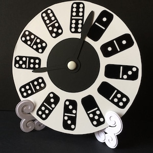Könnte beinhalten: Eine weiße Uhr mit schwarzen Dominosteinen als Ziffern. Die Uhrzeiger sind schwarz und das Zifferblatt der Uhr ist in der Mitte schwarz. Die Uhr steht auf einem weißen Ständer mit zwei Spiralen.