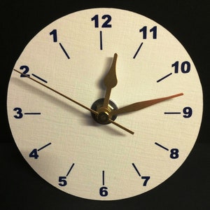 Peut inclure: Une horloge analogique blanche avec des chiffres noirs et des aiguilles dorées. Le cadran de l'horloge a une surface texturée et les aiguilles pointent vers 10h55.