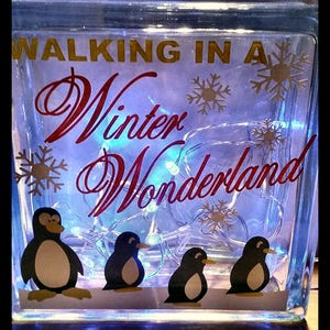 May include: A glass block with a winter wonderland theme. The block features the text "Walking in a Winter Wonderland" with penguins and snowflakes. The block is lit from within with white lights.