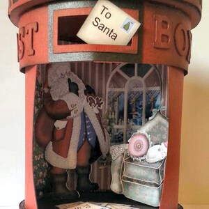 May include: A red and black cardboard mailbox with a cutout window. The window has a white card that says "To Santa" inside. The mailbox has a small Santa Claus figure inside with a white cat sitting in a chair.