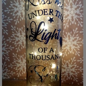 May include: A clear glass bottle with a string of lights inside. The bottle has a black vinyl decal that reads "Kiss me under the light of a thousand stars".