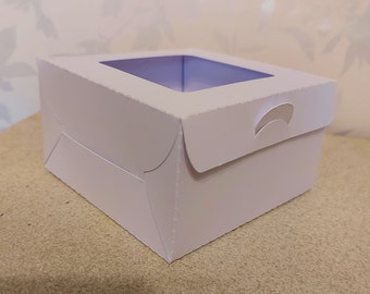 May include: White cardboard box with a clear window on the top. The box has a flap on the top that can be closed.