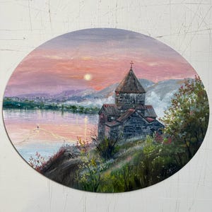 May include: Oval painting of a church on a hill overlooking a body of water at sunset. The sky is a blend of pinks and oranges, with a bright sun. The church is made of stone, and the landscape includes trees and mountains.