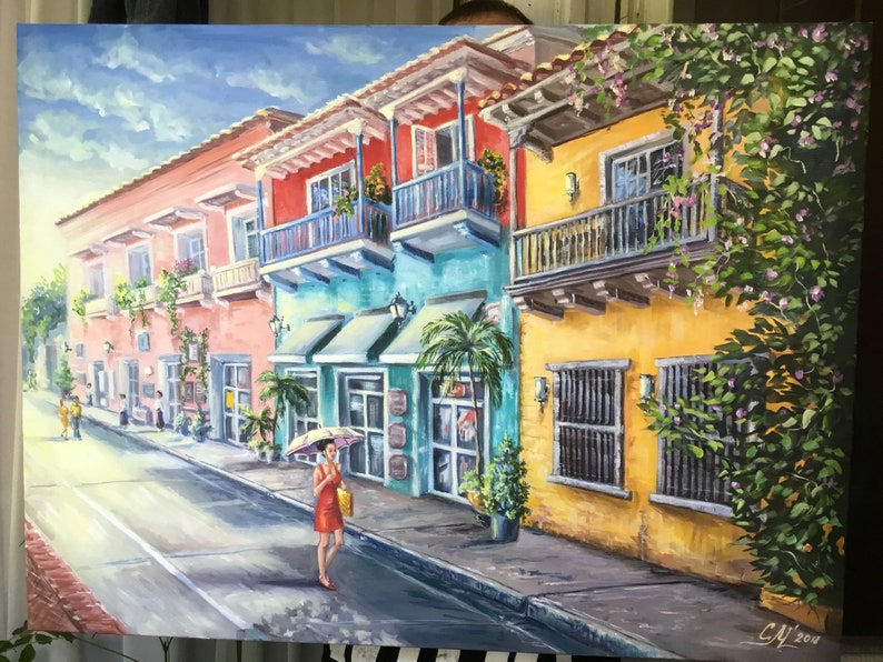 May include: A painting of a colorful street scene in a tropical city. The buildings are painted in shades of yellow, pink, blue, and green. There are balconies with railings and lush green plants growing on the walls. A woman in a red dress walks down the street with an umbrella.