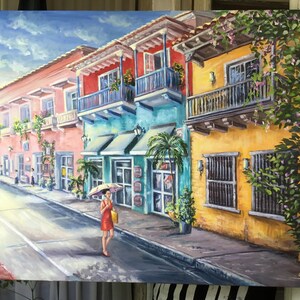 May include: A painting of a colorful street scene in a tropical city. The buildings are painted in shades of pink, yellow, blue, and green. There are balconies with wrought iron railings and lush green plants growing on the walls. A woman in a red dress walks down the street with a yellow umbrella. The sky is blue with white clouds.