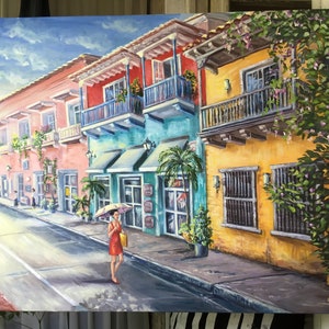 May include: A painting of a colorful street scene in a tropical city. The buildings are painted in shades of pink, yellow, blue, and green. There are balconies with plants and flowers. A woman in a red dress walks down the street with an umbrella. The painting is signed "C.M. 2014".