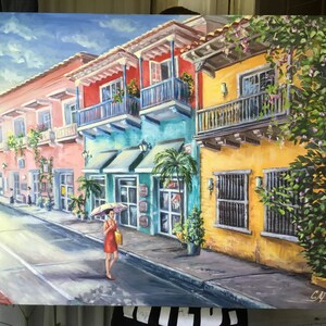 May include: A colorful painting of a street scene in a tropical city. The buildings are painted in shades of pink, yellow, and blue, and have balconies with ornate railings. There are palm trees and lush greenery in the foreground, and a woman in a red dress is walking down the street with an umbrella.