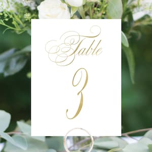 May include: White card with gold calligraphy that reads "Table 3" on a gold metal stand. The card is sitting on a white table with white flowers and green leaves in the background.