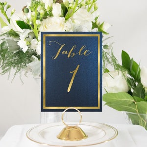 May include: Gold table number card with the text "Table 1" on a navy blue background. The card is sitting on a gold stand.
