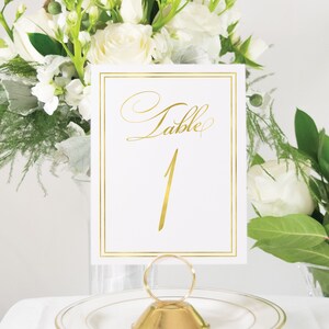 May include: Gold table number card with a white background and a gold border. The card says "Table 1" in a gold script font.