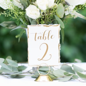 May include: White table number card with gold lettering that says "table 2" on a gold metal stand. The card has a distressed, torn edge look. The stand is on a white tablecloth.