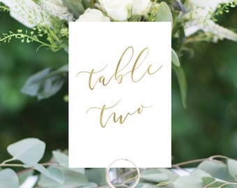 May include: White card with gold calligraphy that reads "table two" on a gold metal stand. The card is sitting on a white table with green foliage.