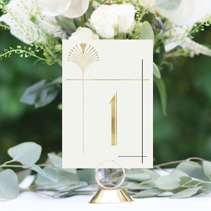 May include: A cream-colored table number card with gold art deco design and the number 1. The card is held in a gold metal holder. White flowers and greenery are in the background, creating an elegant aesthetic.