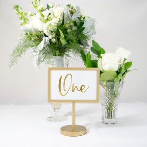 May include: Gold table number holder with a white card that says "One" in gold script. The holder is on a white table with white flowers in vases.
