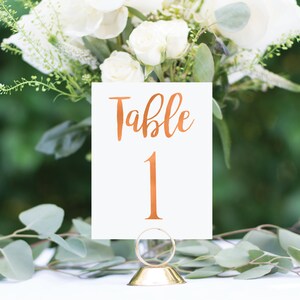 May include: A white card with the words "Table 1" written in copper foil lettering. The card is on a gold metal stand and is surrounded by greenery.