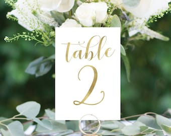 May include: White card with gold lettering that reads "table 2". The card is on a gold stand and surrounded by white flowers and green leaves.