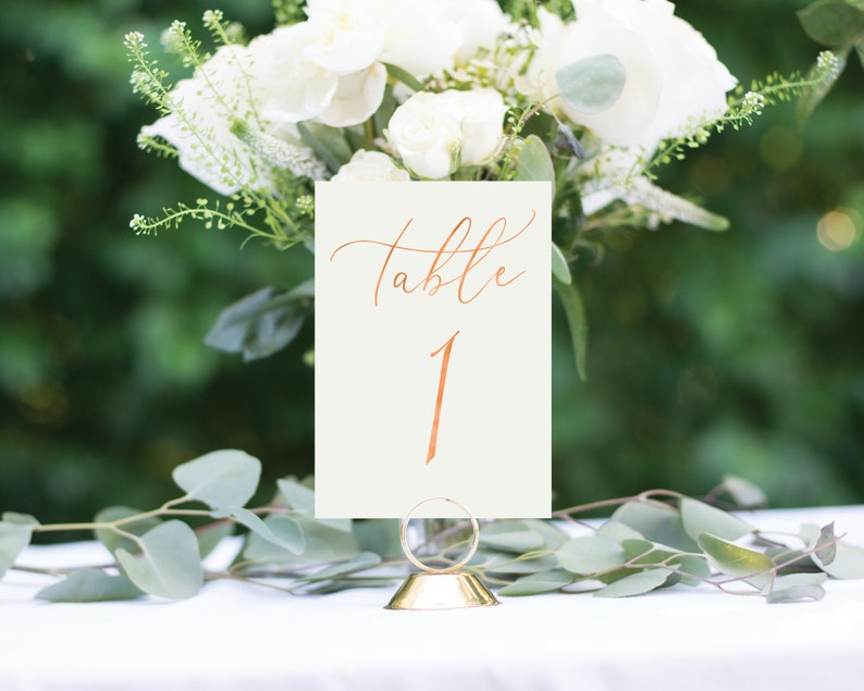 May include: A white card with the words "Table 1" written in copper-colored script. The card is on a gold metal stand and is surrounded by white flowers.