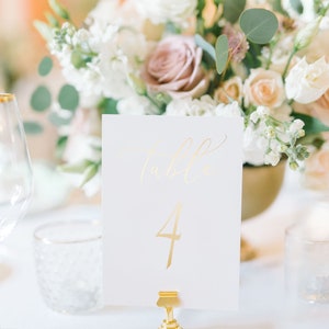 May include: Gold table number card with the number "4" written in gold script. The card is on a gold stand and is in front of a white floral arrangement.