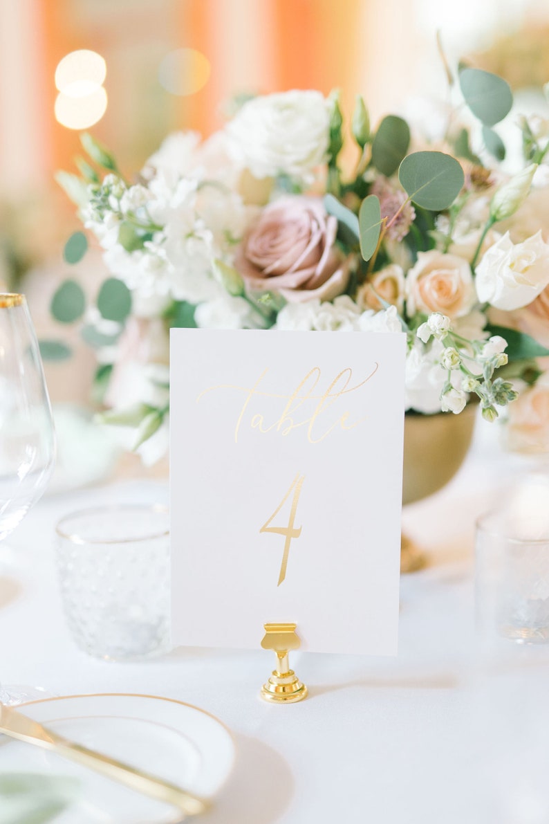 May include: Gold table number card with the number "4" written in gold script. The card is on a gold stand and is in front of a white floral arrangement.