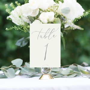 May include: A white card with the text "Table 1" in gray calligraphy, displayed on a gold metal stand. The card is surrounded by white flowers and green foliage.