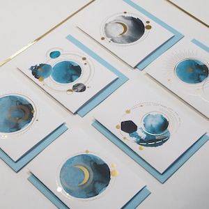May include: Set of six greeting cards with blue and gold watercolor designs featuring moons and stars. The cards have a white background and blue envelopes.