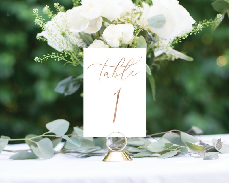 May include: White card with gold calligraphy that reads "Table 1" on a gold metal stand. The card is sitting on a white table with green foliage in the background.