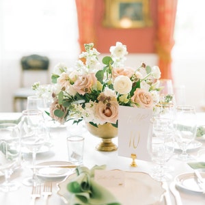 May include: A white table setting with a gold rimmed plate, green napkin, and gold silverware. A centerpiece of pink and white flowers sits in a gold vase. A white card with gold lettering reads "Table 10".