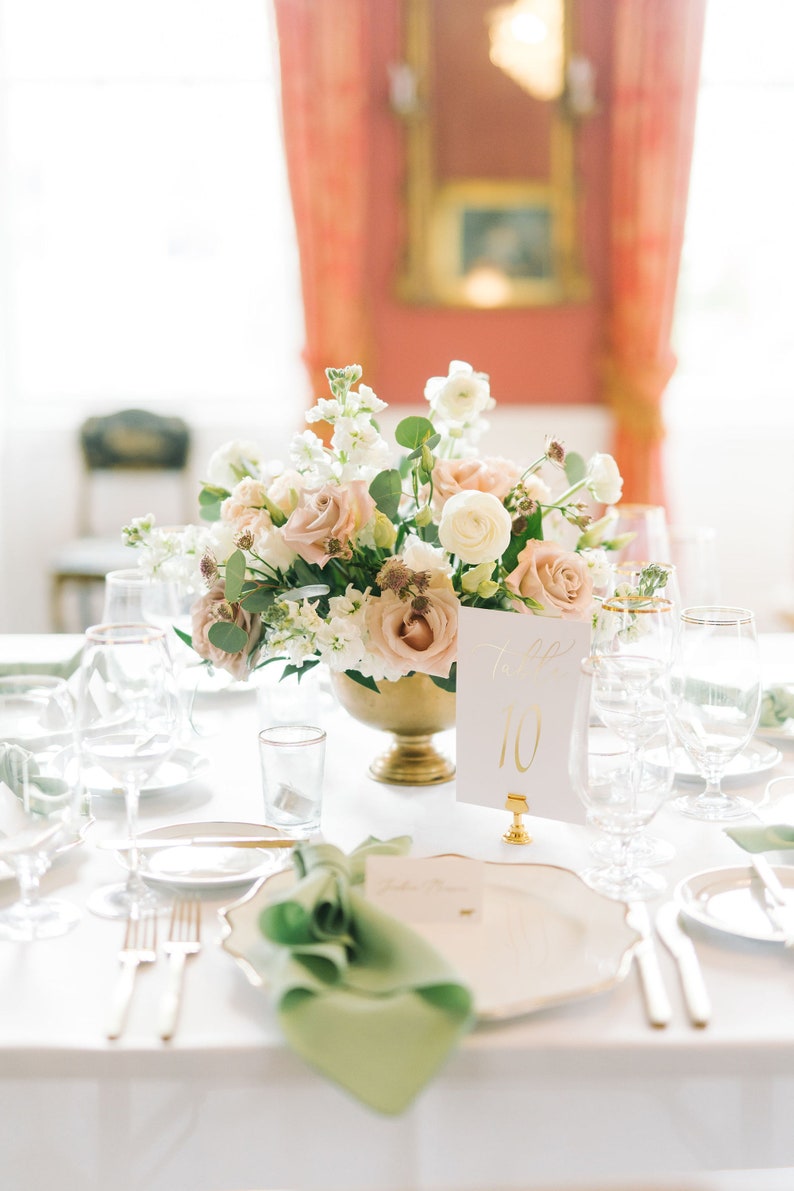 May include: A white table setting with a gold rimmed plate, green napkin, and gold silverware. A centerpiece of pink and white flowers sits in a gold vase. A white card with gold lettering reads "Table 10".
