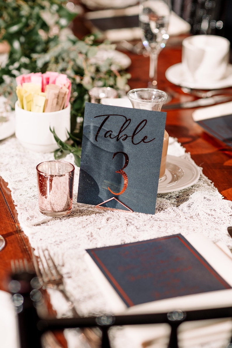 Puede incluir: Una tarjeta de n&uacute;mero de mesa azul oscuro con el texto "Mesa 3" en purpurina de cobre. La tarjeta est&aacute; sobre un camino de mesa de encaje blanco y una mesa de madera.