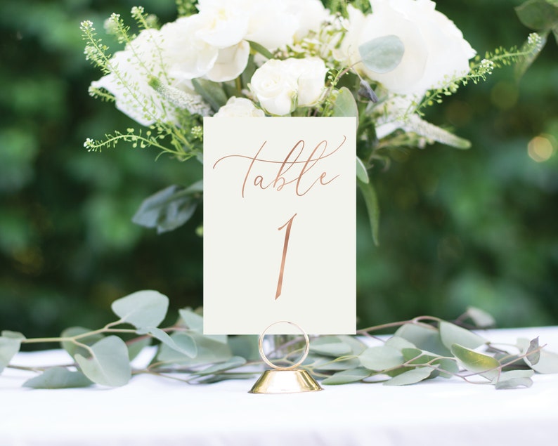May include: A white card with the words "Table 1" written in gold calligraphy. The card is on a gold metal stand and is surrounded by white flowers.