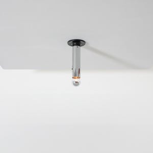 May include: A modern, chrome-finished pendant light fixture with a black circular base. The light has a cylindrical body with a small, spherical bulb at the bottom. The fixture is suspended from a white ceiling.