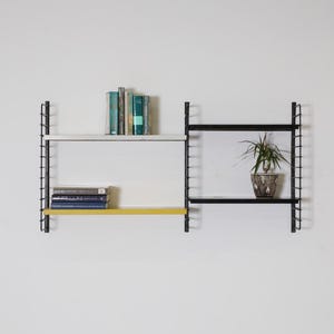 May include: A modular wall-mounted shelving unit with black metal supports and shelves in white, yellow, and black. Books and a potted plant are displayed on the shelves. The unit is mounted on a white wall.