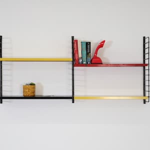 May include: A black metal wall-mounted shelving unit with three shelves. The top shelf is red, the middle shelf is yellow, and the bottom shelf is black. The shelves hold books, a red telephone, and a small plant in a woven pot.