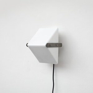 May include: A white, geometric wall-mounted light fixture with a black cord. The fixture is attached to the wall with a metal bracket. The design is minimalist and modern, suitable for interior lighting.