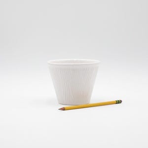 May include: A white, textured ceramic cup with a pencil. The cup has a tapered shape and a ridged pattern. The yellow pencil with a pink eraser is approximately 7.5 inches long.
