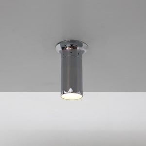 May include: A cylindrical, chrome-finished ceiling light fixture. The light has a rounded top and a narrow, elongated body. The bottom of the light fixture emits a warm, white glow. The fixture is mounted on a white ceiling.