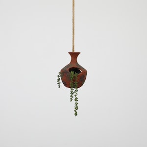 May include: A hanging, terracotta-colored ceramic planter with a textured surface. The planter has a round opening and is suspended by a natural rope. Trailing green plants cascade from the opening, creating a decorative display.