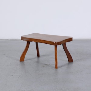 May include: A small, wooden stool with a curved top and four legs. The stool is made of a warm-toned wood and has a simple, elegant design. The legs are curved, adding a touch of visual interest.