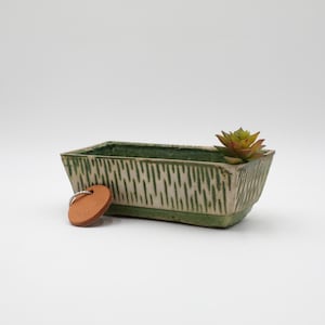 May include: A rectangular ceramic planter with a green and cream textured design, holding a small succulent. A small, round, brown leather tag with a silver ring is next to the planter.
