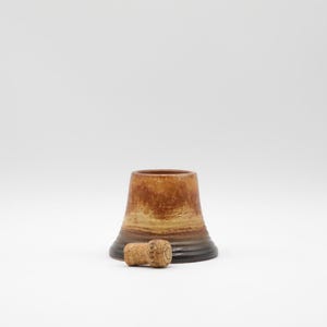 May include: A brown and tan bottle stopper with a cork. The stopper has a flared shape with a dark brown base and a lighter brown top. The cork is cylindrical and lies next to the stopper.