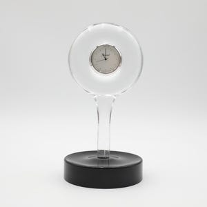 May include: A clear glass desk clock with a circular face and a silver-tone dial. The clock is supported by a clear stem and a black, circular base. The clock's design is minimalist and modern.