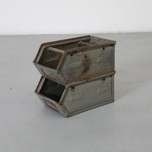 May include: Two stacked, weathered metal storage bins. The bins are a muted green-gray with visible rust and wear. Each bin has an open top and a rectangular shape, with a handle on the top bin.