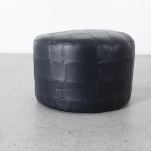 May include: A dark navy blue, round ottoman with a woven, square pattern. The footstool is a solid color and sits on a gray concrete floor. The background is a plain white wall.